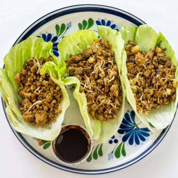 Three Howard Wang's Preston Hollow lettuce wraps filled with ground meat and a side of sauce.